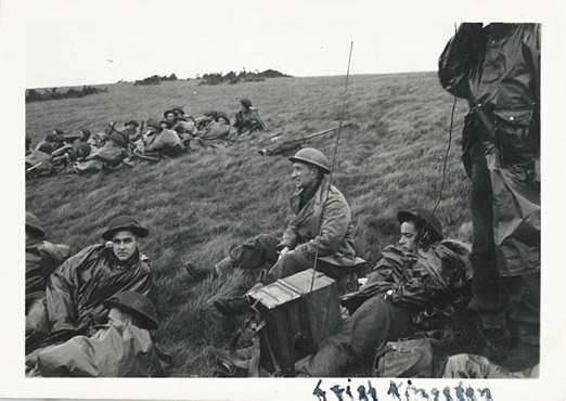 Photographie en noir et blanc – Archie, en tenue militaire complète, est assis dans l’herbe, entouré d’autres soldats au repos. Il ne regarde pas la caméra. Les mots écrits dans un coin de la photo sont flous.